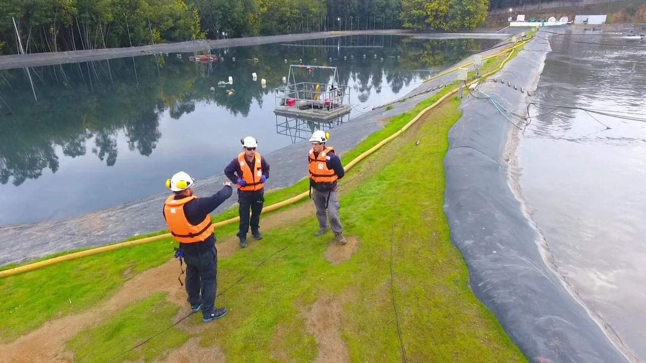 Trabajadores con casco y chaleco realizando un análisis seguro de trabajo junto a una laguna de proceso