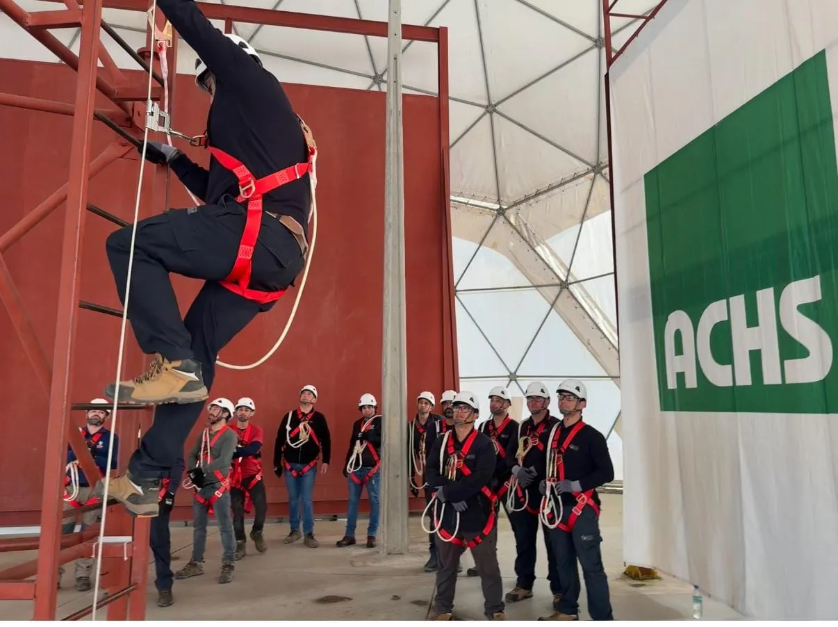 Curso práctico de trabajo en altura: trabajador con arnés suspendido de una línea de vida frente a un grupo de alumnos, con pendón de ACHS